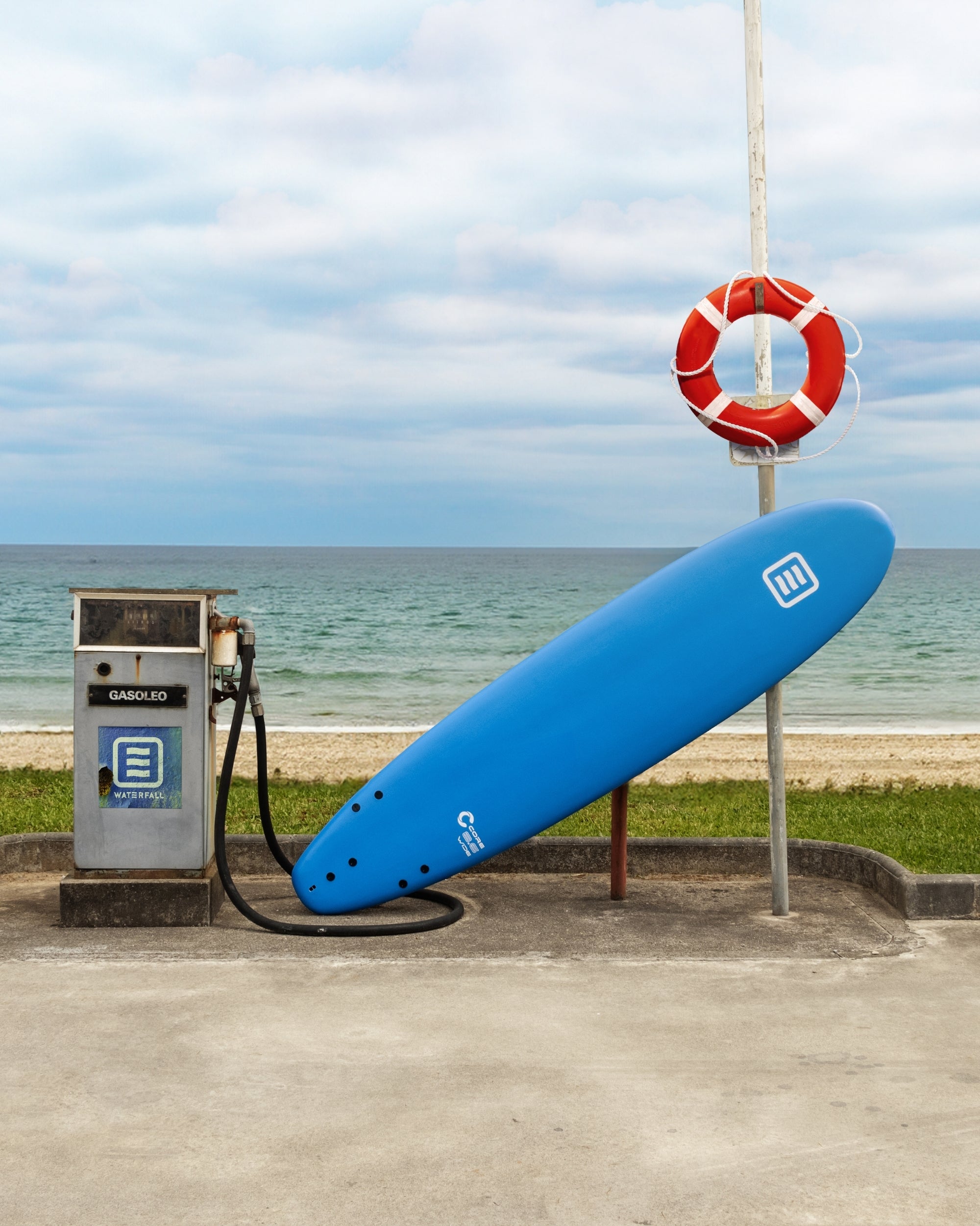 Blue surfboard core with 8" size, shown on a stand near the water.